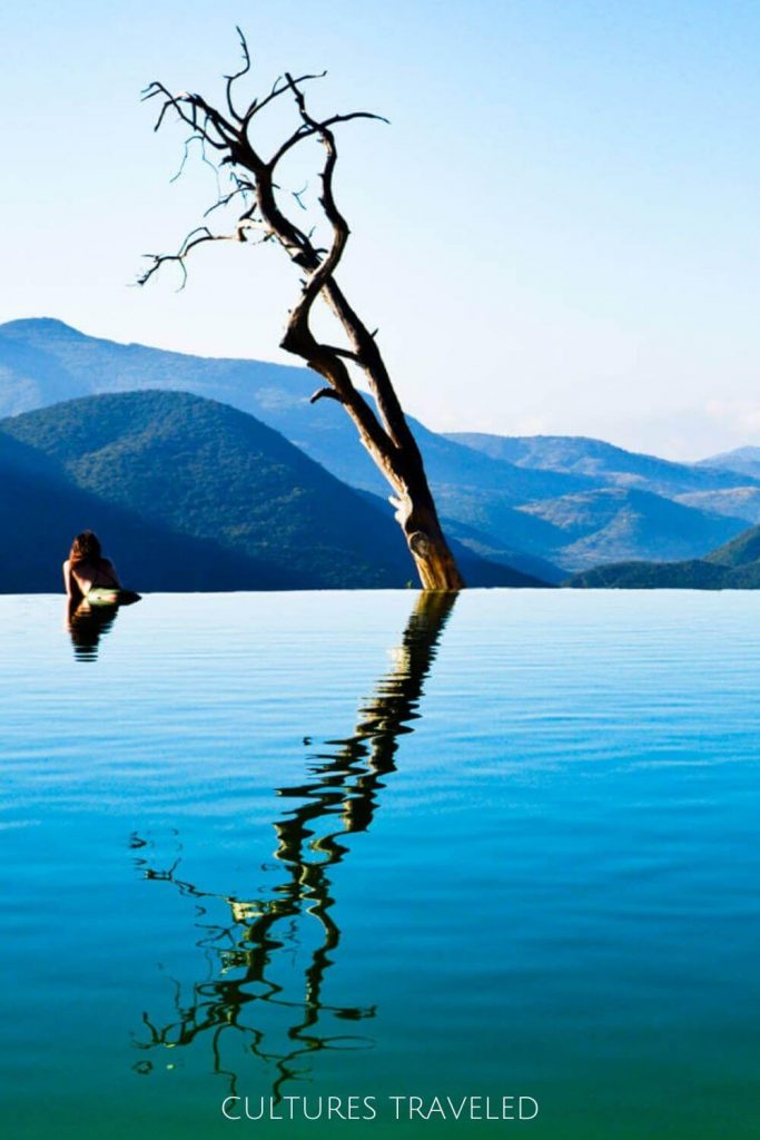 A woman looks over the mountains as she rests at the edge of the infinity pool at Hierve el Agua with the large leafless tree to her right reflecting in the pool.