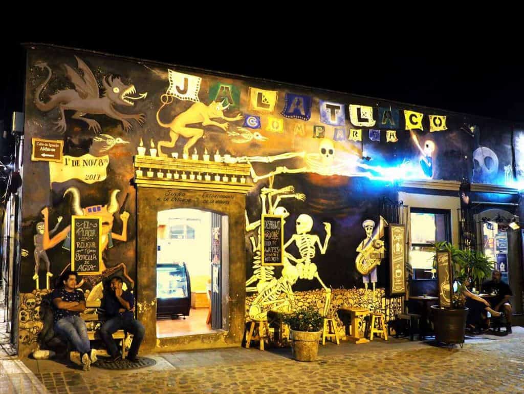 Two men sit outside of a restaurant in Jalatlaco, Oaxaca at night. The front of the restaurant is painted with a vibrant day of the dead murals that includes skeletons, candles, and mythical figures.